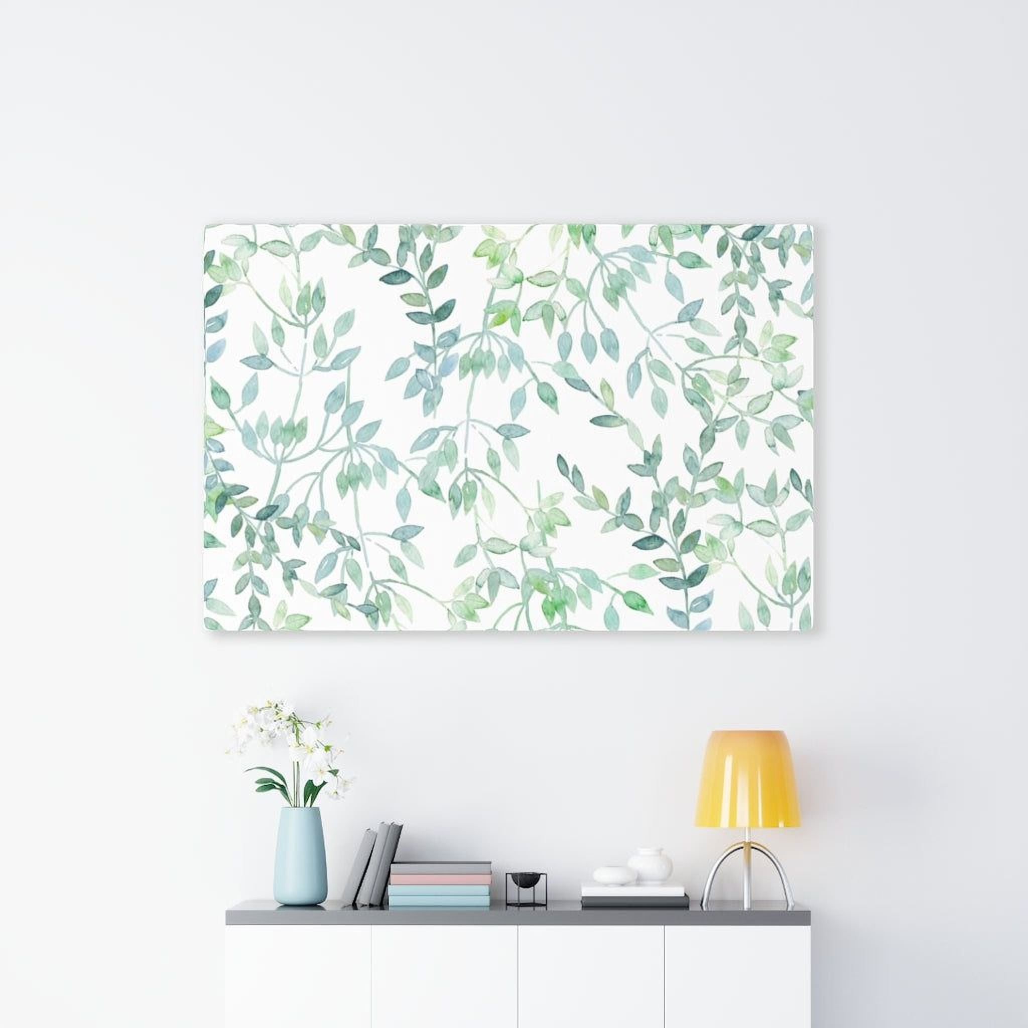a white wall with a framed print of green leaves hanging above a shelf with a vase of white flowers, a yellow lamp, and a stack of books.