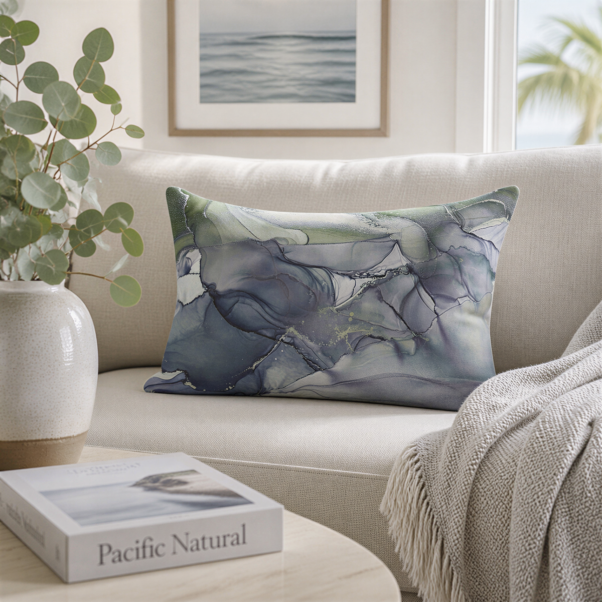 a living room with a white sofa, a decorative pillow with a marble-like pattern, a white vase with greenery, a book titled "Pacific Natural", and a framed picture on the wall.
