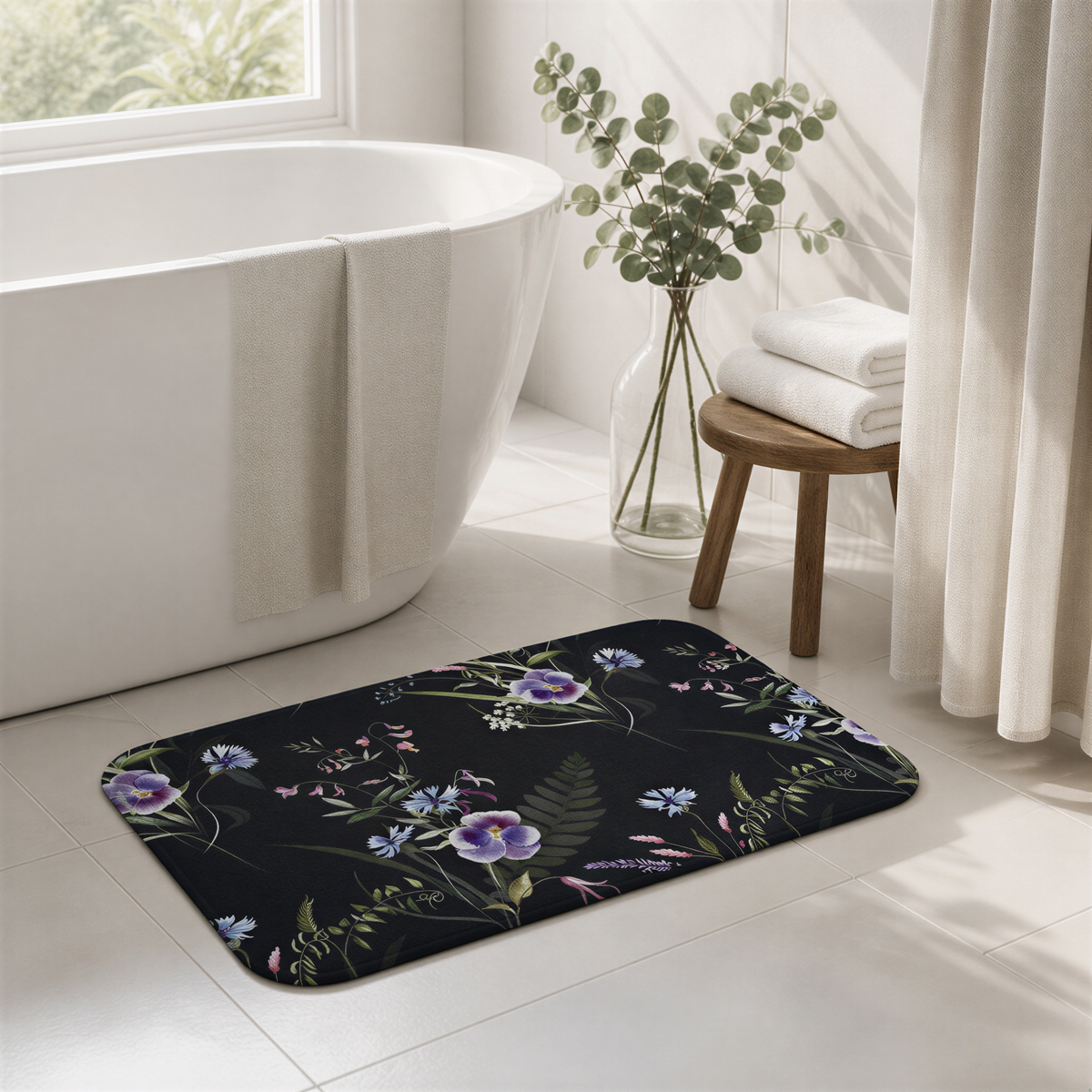a bathroom with a white bathtub, a black floral bath mat, and a vase of greenery on a wooden stool.