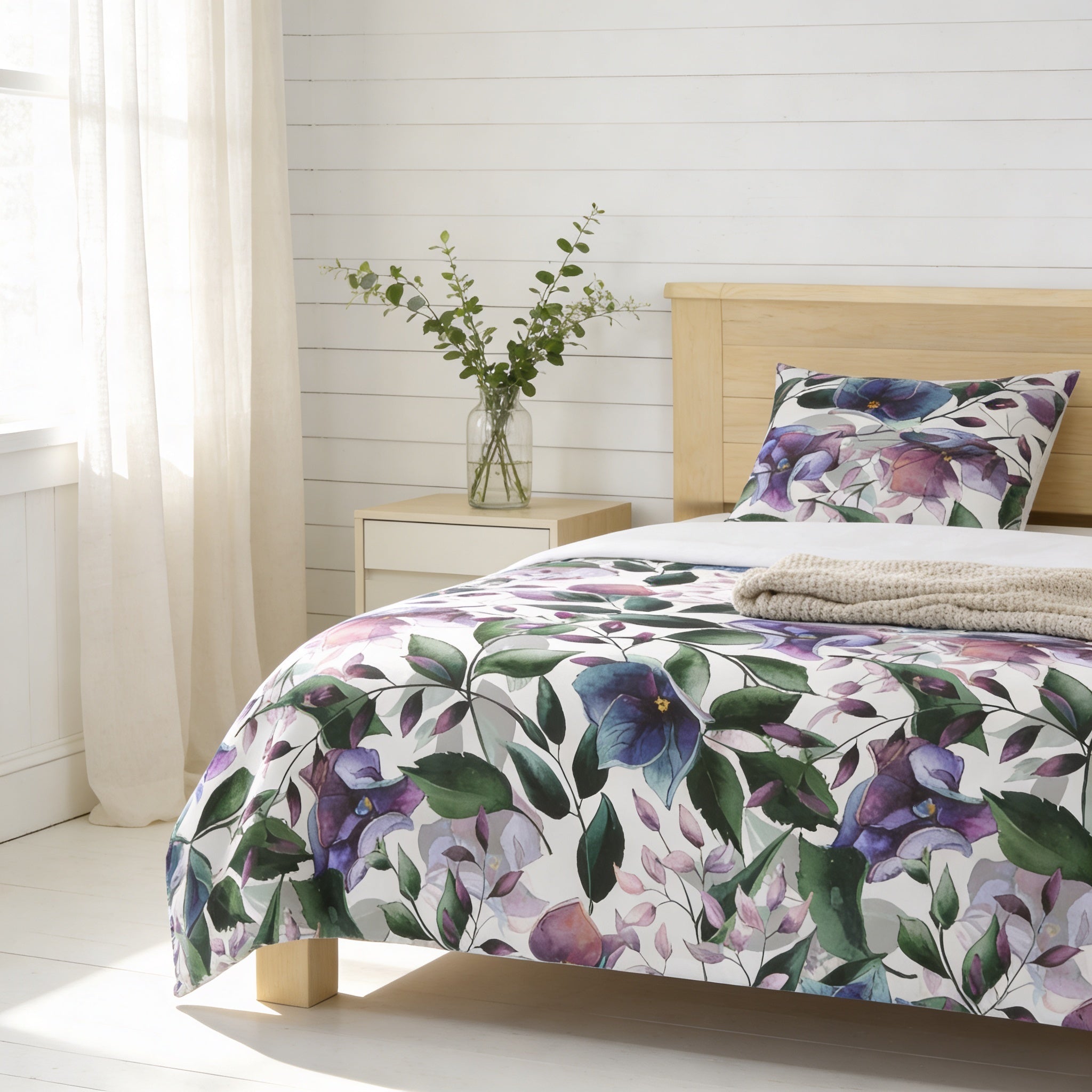 a bedroom with a floral bedspread, a wooden headboard, and a small table with a vase of greenery.