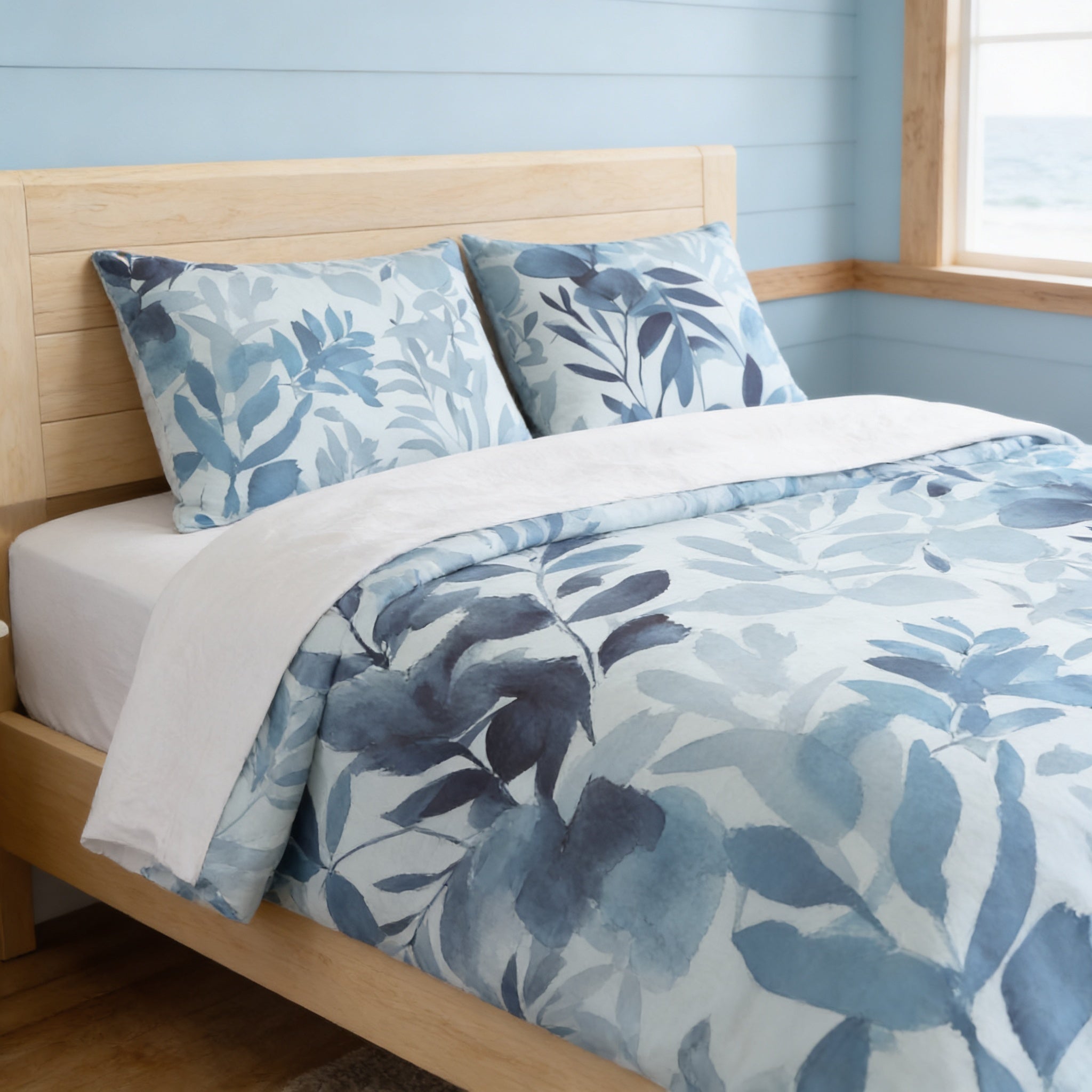 a bedroom with a bed featuring a blue and white leaf-patterned comforter and pillows. The bed is set against a light blue wall, and there is a window nearby.