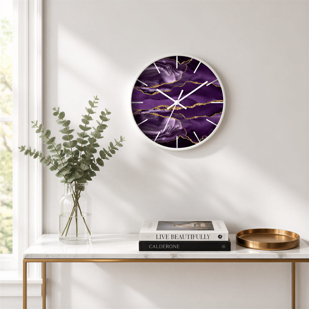 a white wall with a round clock displaying a purple marble design, a vase of greenery, a stack of books, and a gold tray on a white shelf.