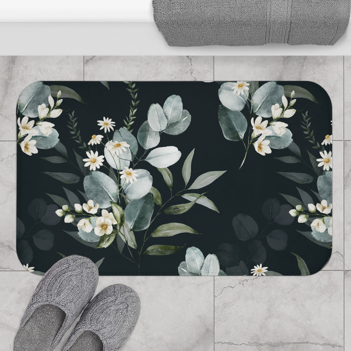 A bathroom mat with a floral design, featuring white flowers and green leaves against a dark background. The mat is placed on a tiled floor, with a pair of gray slippers visible underneath it.