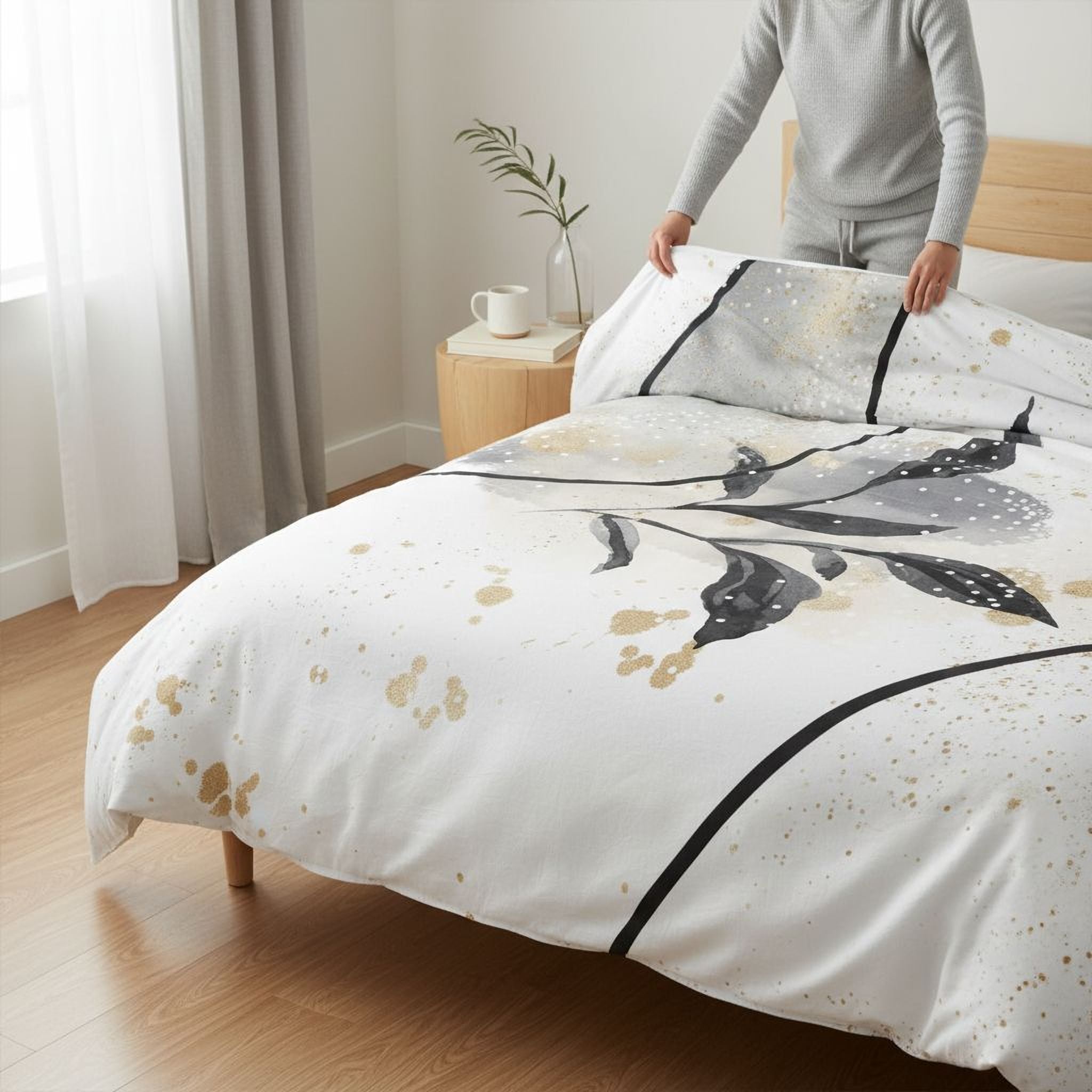 A person is arranging a bed with a white and black patterned comforter, which features a bird design. The bed is set against a backdrop of a wooden headboard and a window with white curtains.