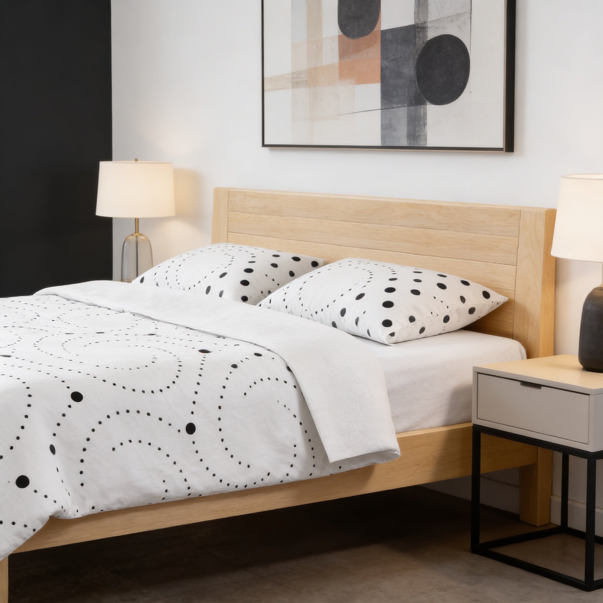 a modern bedroom with a wooden bed frame, white bedding with black polka dots, and two nightstands with lamps. The room has a minimalist and clean design, with a large abstract painting on the wall above the bed.