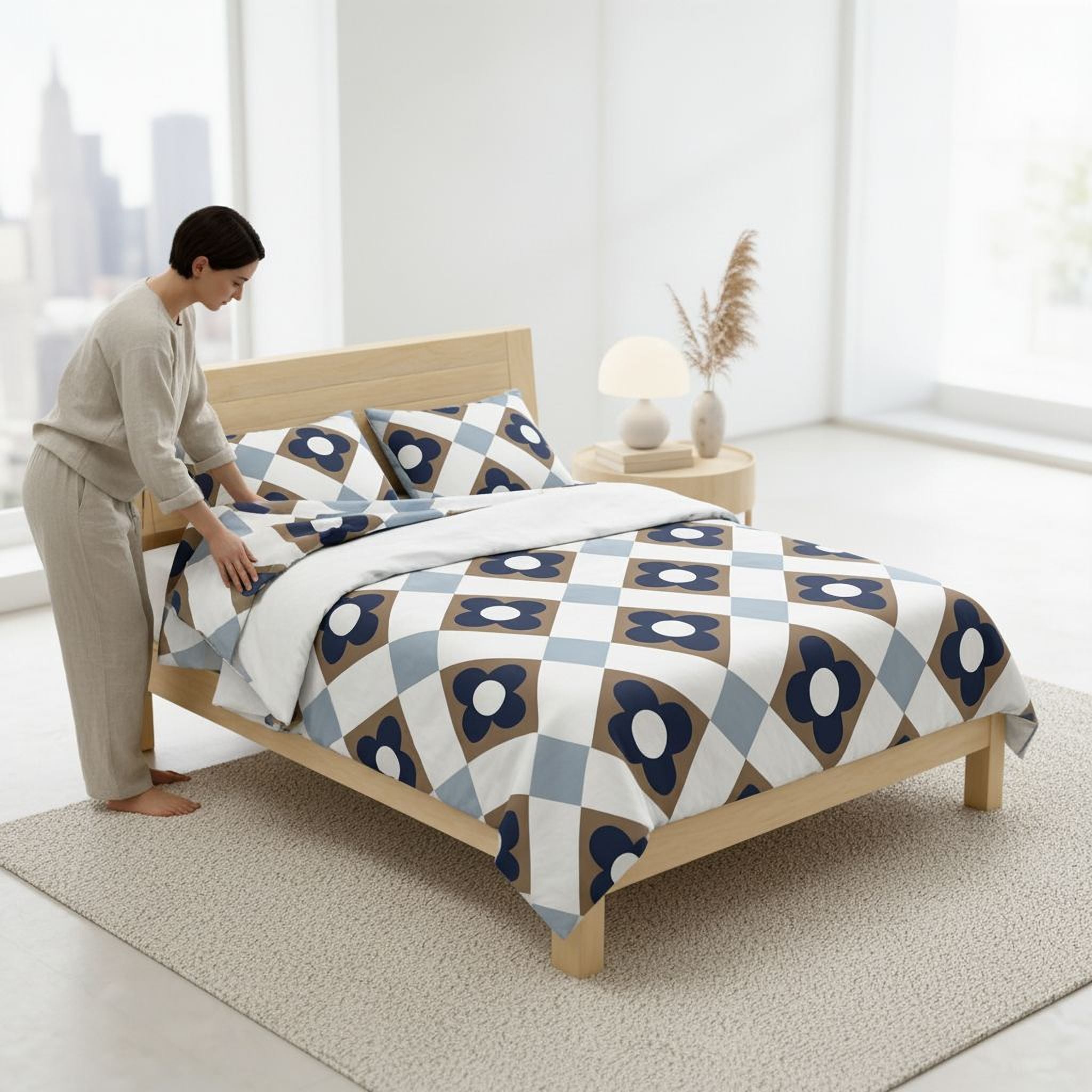 A woman is arranging a bed with a blue and white patterned comforter and pillows in a bedroom with a large window overlooking a city skyline.