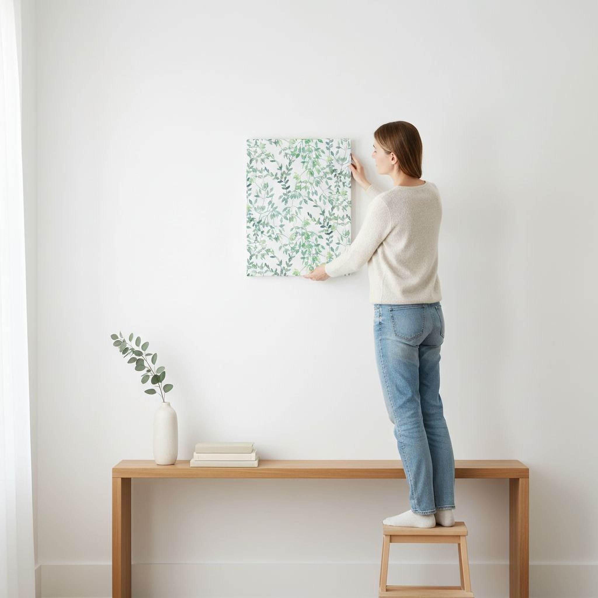 A woman is standing on a stool and hanging a framed picture on a white wall.