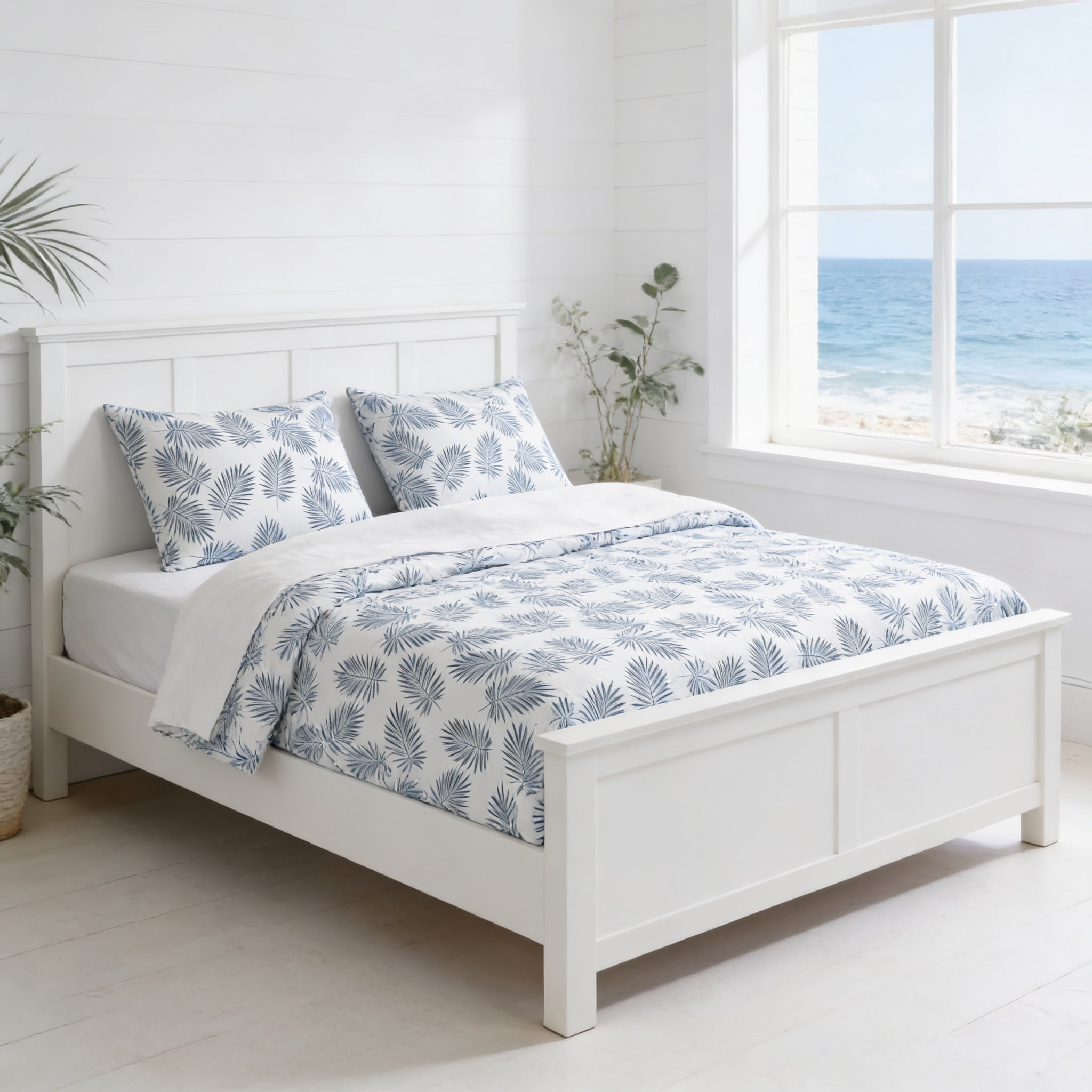 A white wooden bed with a blue and white patterned comforter and pillows, situated in a bright and airy bedroom with a large window overlooking the ocean.