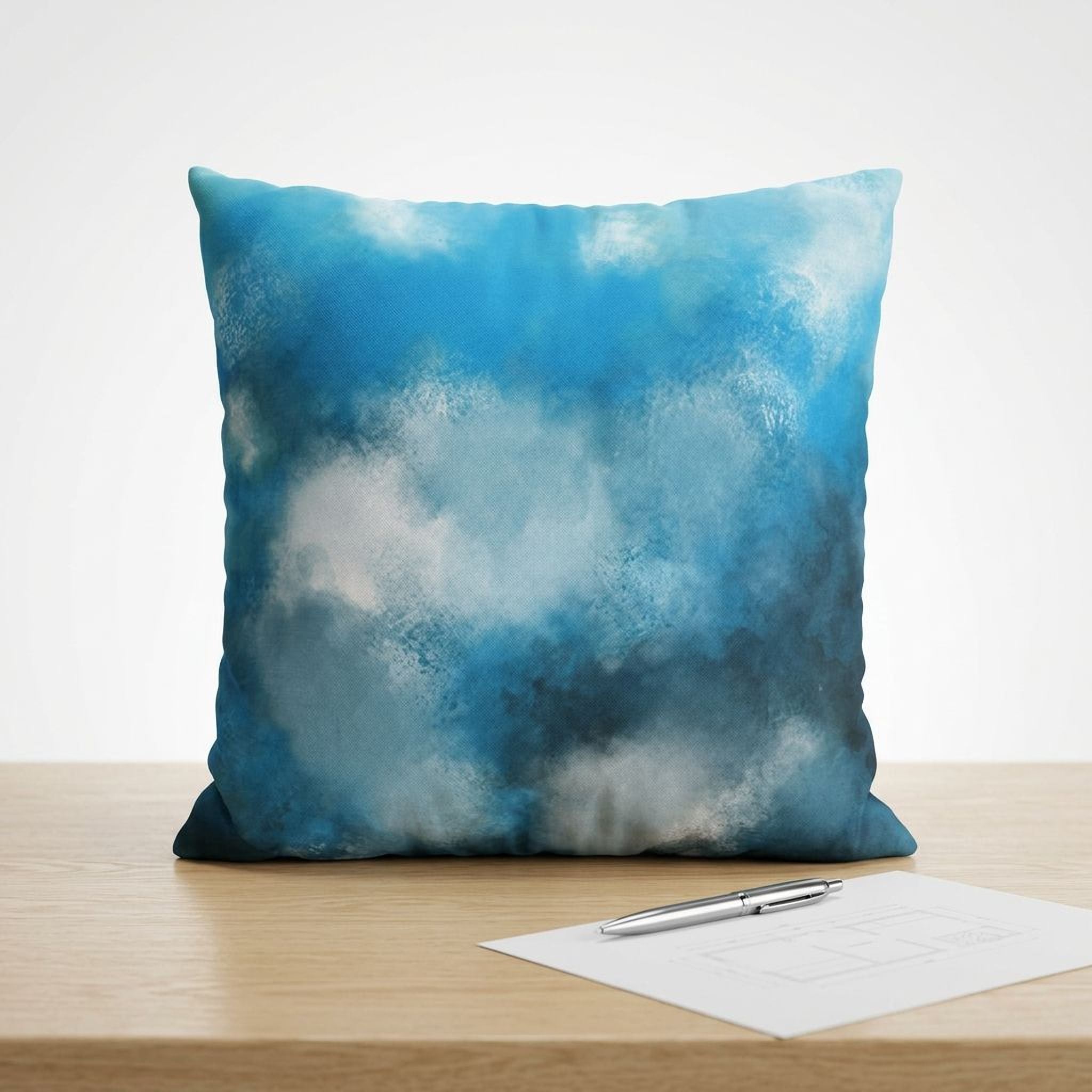 A blue and white cloud-like pattern on a pillow is displayed on a wooden surface, accompanied by a pen and a piece of paper.