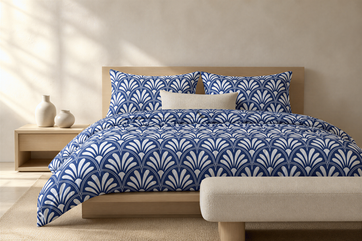 a bedroom with a bed dressed in a blue and white patterned comforter and pillows. The bed is positioned against a wall, with a wooden headboard. There is a wooden nightstand next to the bed, and a white bench is placed in front of it. The room has a minimalist and modern design, with a neutral color palette and clean lines.