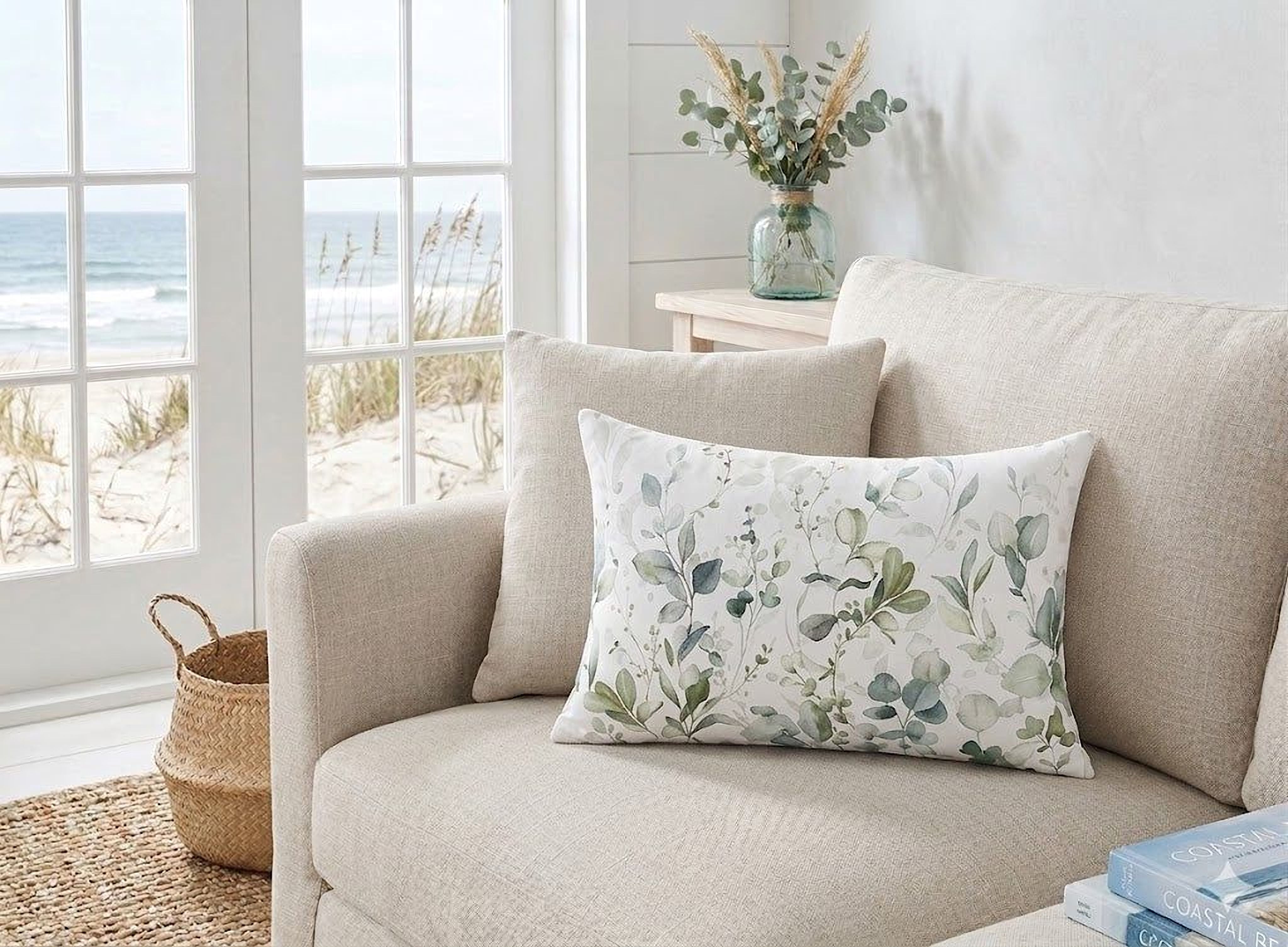 a cozy living room with a beige sofa, a decorative pillow with a floral pattern, and a vase of greenery on a nearby table. The room has large windows that offer a view of the ocean, creating a serene and relaxing atmosphere.