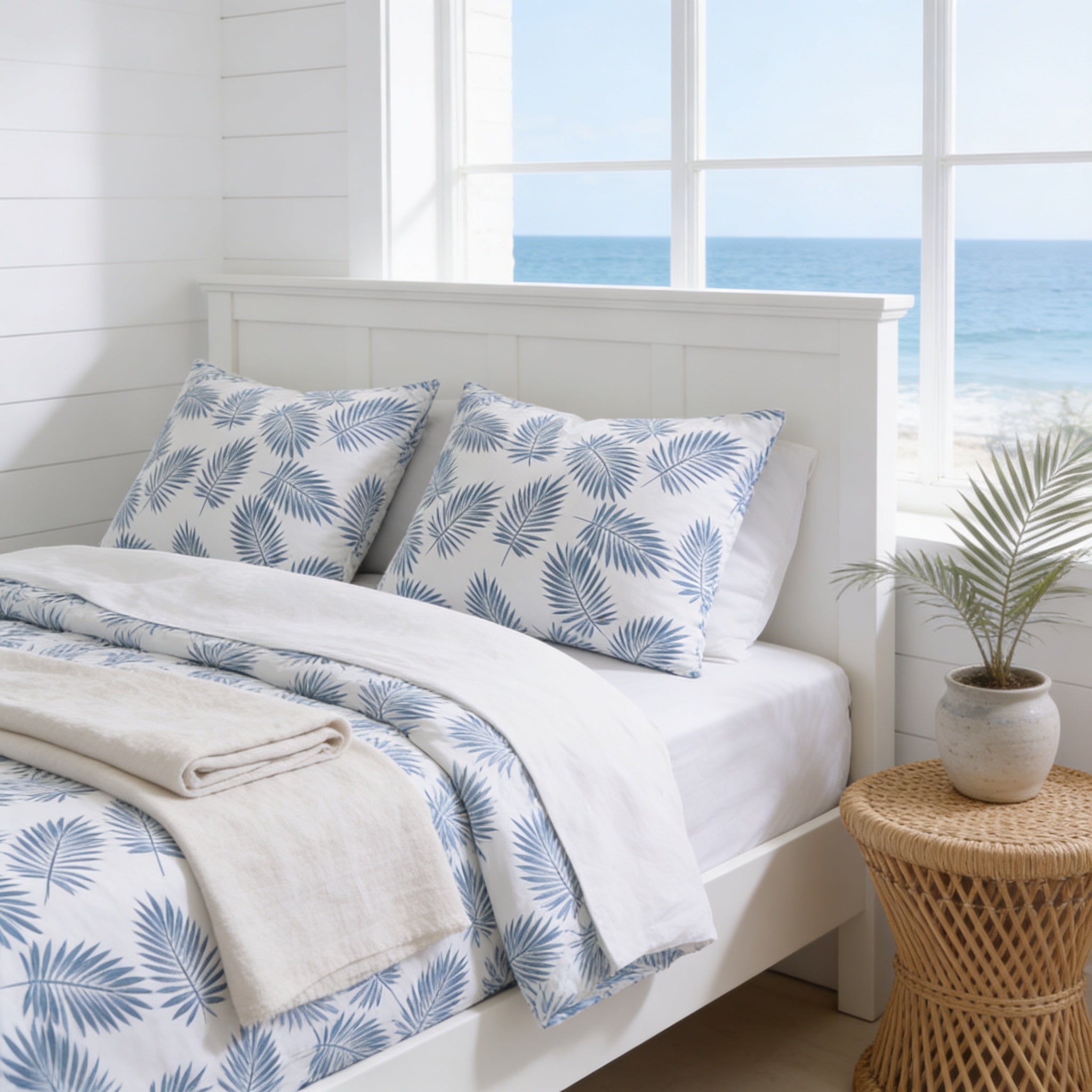 a serene bedroom scene with a white bed frame, a white headboard, and a window overlooking the ocean. The bed is adorned with blue and white patterned bedding, and a woven side table is placed next to the bed. A potted plant is also present in the room, adding a touch of greenery to the space.